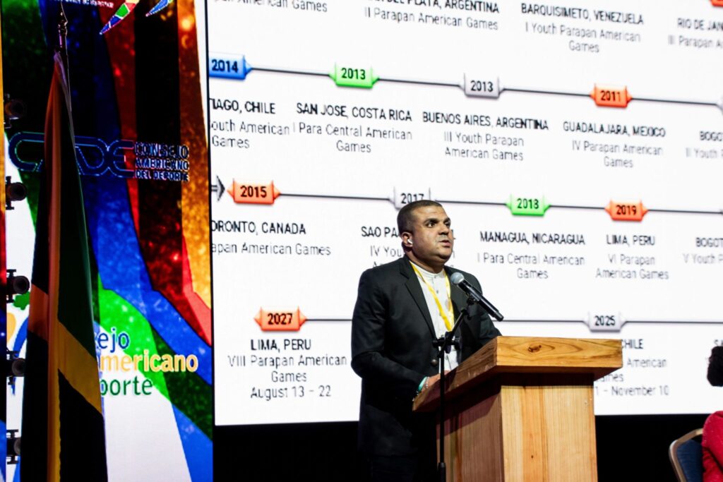 En imagen se observa en el estrado al presidente del AmPC, Julio César Ávila, disertando ante los presentes, vistiendo un traje formal y delante de banderas de protocolo en el escenario.