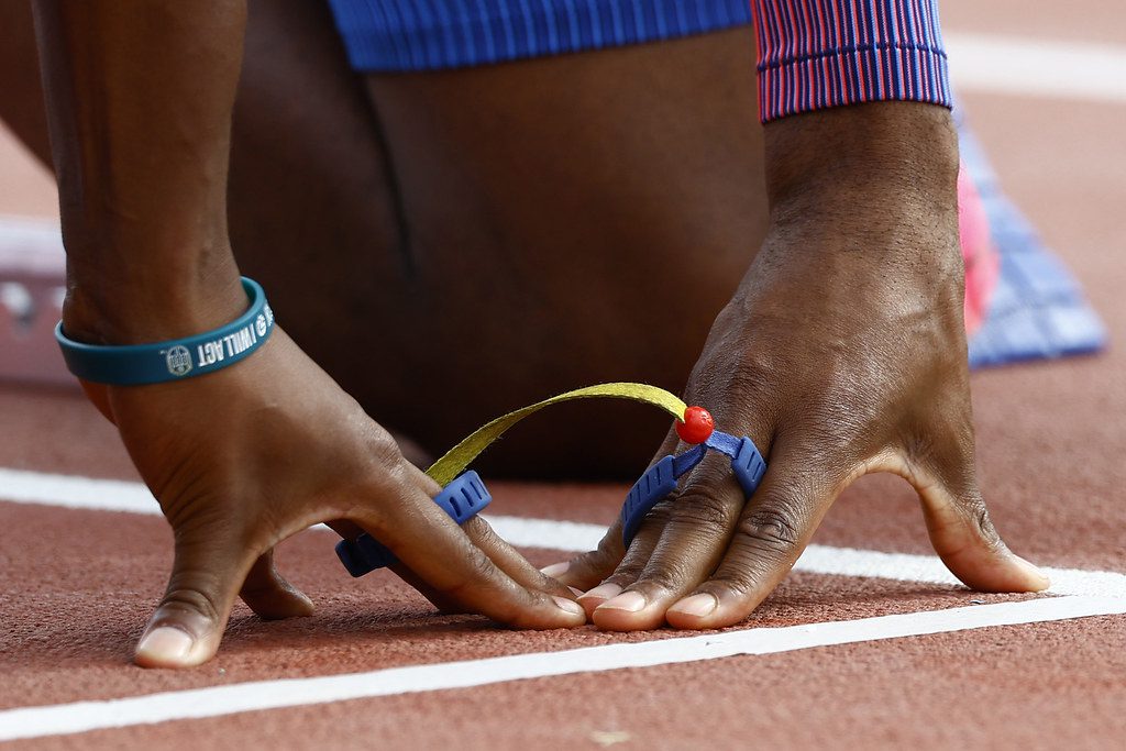 Dos manos de personas distintas, unidas por una especie de cordón. ambos tocan la pista de atletismo como a punto de largar.