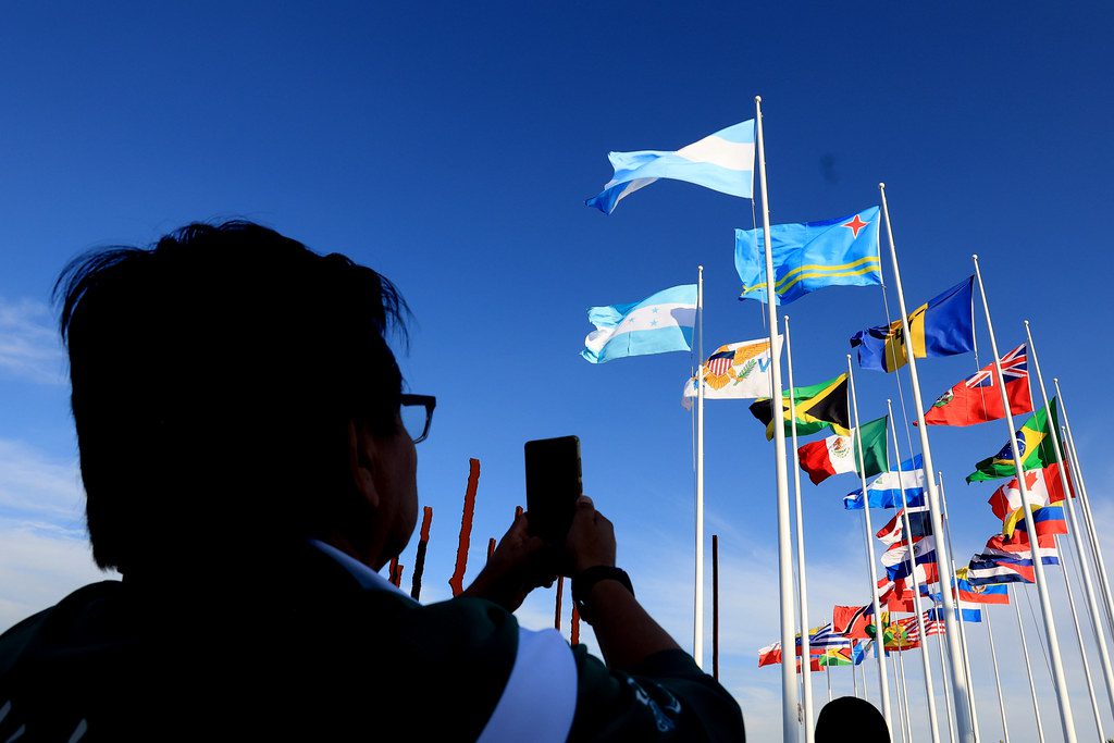 En la foto: una persona a contraluz, tomándole foto a las distintas banderas de los países miembros del AmPC.