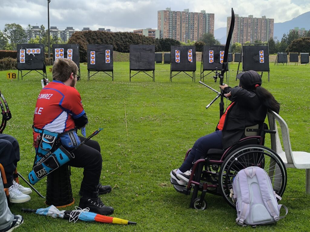 En la foto dos para atletas sentados, uno de ellos en silla de ruedas, apuntando para disparar.