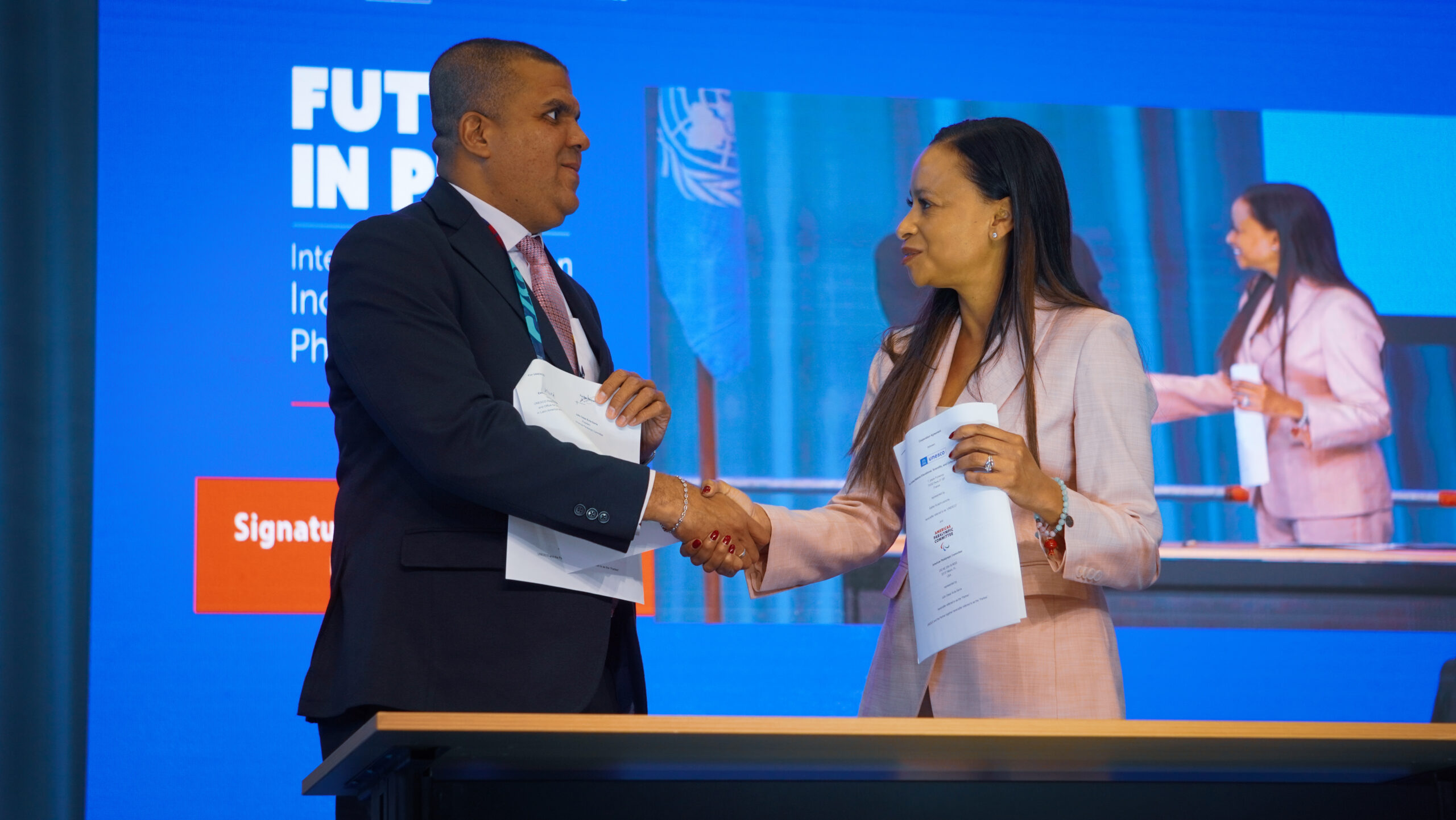 en la foto Julio César Ávila y Esthér de UNESCO, ambos firmando el acuerdo y dándose la mano.